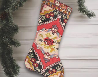 May include: A Christmas stocking with a red, black, and cream floral pattern. The stocking is suspended by a white string, with a Christmas tree branch and decorations in the background. The stocking has a traditional design.
