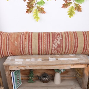May include: A long, rectangular pillow with a woven design in shades of beige, orange, and red. The pillow features vertical stripes and geometric patterns. It rests on a wooden bench, suggesting a rustic or bohemian style.