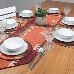 Puede incluir: Una mesa de madera con un mantel individual estampado en rojo, naranja y marrón. La mesa está puesta con platos, cuencos y vasos blancos. Hay dos sillas blancas a cada lado de la mesa.