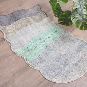 Op de afbeelding: Vijf gelaagde kattenvormige tapijten in verschillende tinten grijs, beige en groen. De tapijten zijn gemaakt van een zacht, geweven materiaal en hebben een verweerde, vintage uitstraling.
