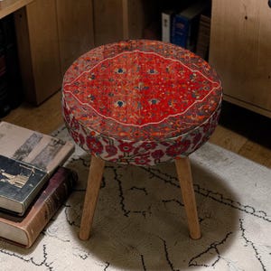 May include: Round ottoman with a red and orange patterned top, featuring a design reminiscent of a rug. The sides display a floral pattern. It has three wooden legs.