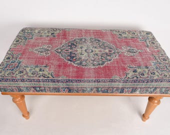 May include: A red, blue, and green patterned ottoman with wooden legs. The ottoman is upholstered with a vintage rug.