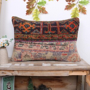 May include: A colorful, rectangular throw pillow with a traditional Turkish kilim pattern. The pillow is made of woven fabric and has a variety of colors, including red, orange, blue, green, and brown. The pillow is sitting on a wooden shelf.