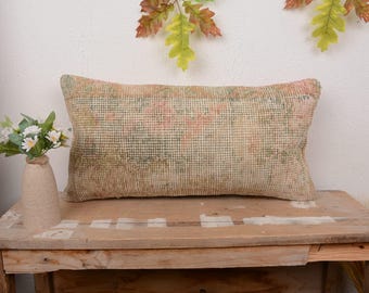 May include: A rectangular cushion in beige with a faded floral pattern in pink and green. It rests on a wooden table. A small vase with white flowers and greenery is to the left.