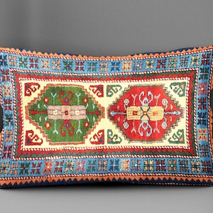May include: Rectangular decorative pillow with a colorful, intricate rug design. The pillow features a central cream-colored field with two patterned medallions in green and red. The border is blue with orange accents, creating a vibrant, eye-catching piece.