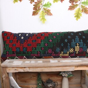 May include: A long, rectangular pillow with a colorful geometric pattern in shades of red, green, blue, and black. The pillow is made of woven fabric and has a fringed edge. It is sitting on a wooden bench.