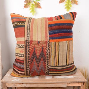 May include: A square patchwork pillow with a variety of patterns and colors. The pillow features a mix of geometric and striped designs in shades of red, orange, blue, and beige. It is placed on a wooden surface.