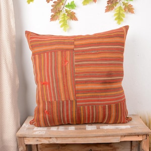 Puede incluir: Un cojín cuadrado con un diseño de patchwork con rayas naranjas, rojas y marrones. El cojín está hecho de tela texturizada y descansa sobre una superficie de madera. Pequeñas costuras de color naranja sirven como detalle decorativo.