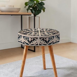 Può includere: Uno sgabello rotondo imbottito con un motivo geometrico beige e blu navy. Lo sgabello ha tre gambe in legno. Lo sgabello è un elemento d'arredo decorativo, adatto per un soggiorno o una camera da letto.