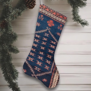 May include: A Christmas stocking with a patterned design in shades of navy blue, rust, and cream. The stocking is hanging from a silver loop against a backdrop of a wooden wall and evergreen branches.