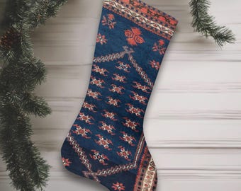 May include: A Christmas stocking with a patterned design in navy blue, rust, and cream. The stocking is suspended by a silver loop against a wooden wall and evergreen branches.