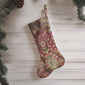 Puede incluir: Un calcetín navideño con un patrón floral y geométrico detallado y multicolor en tonos rojo intenso, verde apagado y dorado. El calcetín tiene un lazo de tela en la parte superior para colgarlo y muestra un diseño textil de estilo tejido.