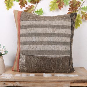 Op de afbeelding: Vierkant decoratief kussen met een gestreept ontwerp in bruin- en beigetinten. Het kussen heeft een patchwork ontwerp en een effen bruine rand. Het kussen ligt op een houten tafel met een vaas bloemen.
