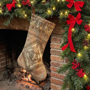 Puede incluir: Un calcetín navideño con un patrón tejido en tonos beige, marrón y crema que recuerda a una alfombra tradicional. El calcetín tiene un lazo simple en la parte superior para colgarlo y muestra un diseño geométrico con bordes intrincados.
