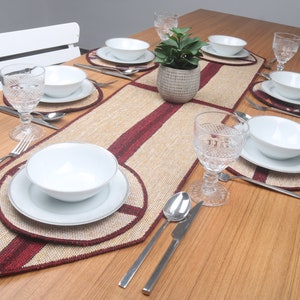Puede incluir: Una mesa puesta con un mantel individual rojo y beige, platos, cuencos y vasos blancos. El mantel individual tiene una textura tejida y una franja roja en el centro. Hay tenedores, cuchillos y cucharas en la mesa.