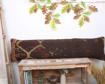 May include: A long brown and red patterned pillow with a geometric design sits on a wooden shelf. The pillow is made of woven fabric and has a fringe on the ends.