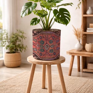 Op de afbeelding: Een groene Monstera-plant met grote, ingesneden bladeren in een cilindervormige stoffen plantenbak. De plantenbak heeft een gedetailleerd, complex patroon in dieprood, zwart en blauwgroen, dat doet denken aan een geweven tapijtontwerp.