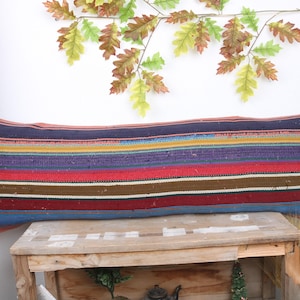 May include: A long, rectangular cushion with a striped pattern in shades of red, blue, purple, brown, and yellow. The cushion is made of woven fabric and has a rustic, handmade look.