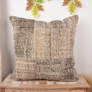 Puede incluir: Cojín decorativo cuadrado con un diseño de patchwork en tonos beige, marrón y gris. El cojín tiene un aspecto texturizado y está colocado sobre una superficie de madera. El cojín es un artículo de decoración del hogar.
