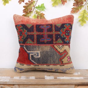 May include: Square decorative pillow with a patchwork design. The pillow features a mix of colors including red, blue, beige, and green. The pillow is placed on a wooden surface, with leaves in the background.