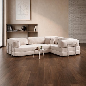 May include: A modern, cream-colored sectional sofa with a low profile and metal accents. The sofa is arranged in an L-shape and sits on a dark wood floor. A small white side table with a plant and a coffee cup is in front of the sofa.
