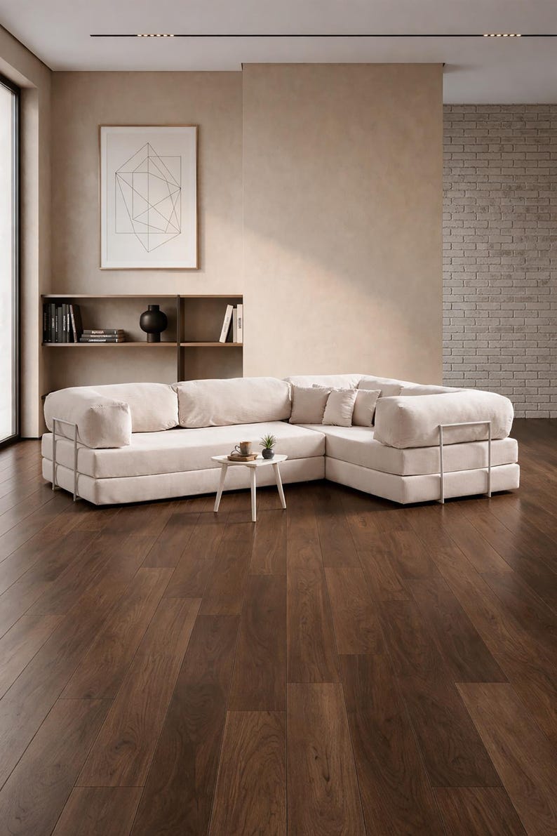 May include: A modern, cream-colored sectional sofa with a low profile and metal accents. The sofa is arranged in an L-shape and sits on a dark wood floor. A small white side table with a plant and a coffee cup is in front of the sofa.