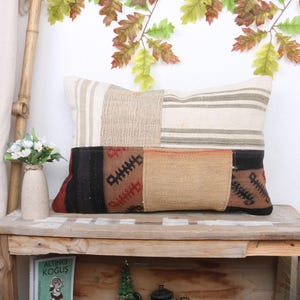 Può includere: Un cuscino rettangolare in patchwork con un tessuto a righe beige e bianco nella parte superiore e un tessuto a motivi marroni, neri e beige nella parte inferiore. Il cuscino è appoggiato su uno scaffale di legno.