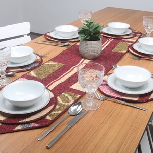 Puede incluir: Un camino de mesa rojo, beige y verde con un patrón geométrico. El camino de mesa está colocado sobre una mesa de madera con seis cubiertos, cada uno con un tazón blanco, un plato blanco y un vaso. Una pequeña planta en maceta está en el centro de la mesa.