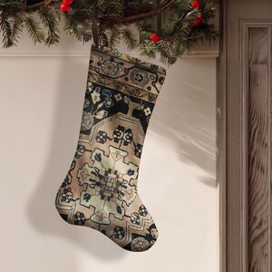 Puede incluir: Un calcetín navideño con un patrón detallado e intrincado en tonos beige, negro y azul oscuro. El diseño imita una alfombra tejida con formas geométricas, motivos florales y bordes simétricos, terminado con una sencilla presilla negra para colgar.