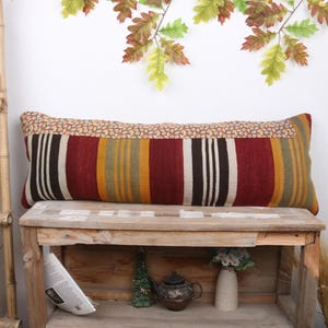 Peut inclure: Un long coussin rectangulaire avec un motif rayé coloré dans des tons de rouge, jaune, marron et blanc. Le coussin est posé sur un banc en bois avec une étagère en dessous.