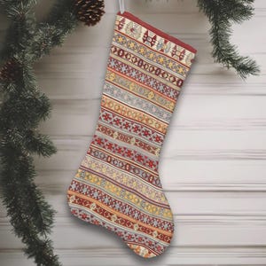 Könnte beinhalten: Ein Weihnachtsstrumpf mit einem gemusterten Design in Rot, Gelb und Beige. Das Design zeigt horizontale Streifen mit geometrischen Formen. Der Strumpf hängt an einem weißen Band.