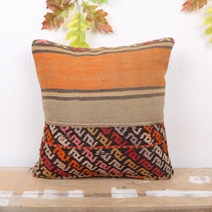 May include: Square decorative pillow with a geometric pattern. The top section features orange and brown horizontal stripes. The middle section is a solid tan color, and the bottom section has a repeating pattern in red, black, and pink.