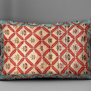 Puede incluir: Cojín decorativo rectangular con diseño de alfombra vintage. El cojín presenta un fondo beige con un patrón de diamantes rojos y detalles florales. Un borde turquesa enmarca el diseño.