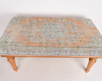 May include: A vintage-style ottoman with a floral patterned fabric top and wooden legs. The fabric is a light peach color with a faded blue and white floral design.