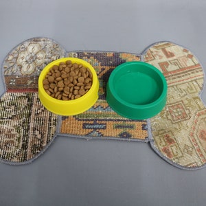 May include: A bone-shaped pet feeding mat with two bowls. One yellow bowl is filled with brown pet food, and the other is a green bowl. The mat is made of a patchwork of patterned fabric.