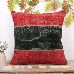 Op de afbeelding: Vierkant decoratief kussen in rood en zwart. Het kussen heeft een rode boven- en onderkant met een zwart middengedeelte. Het zwarte gedeelte heeft een vervaagd, patroonontwerp. Het kussen ligt op een houten oppervlak met een kleine bloemenvaas.