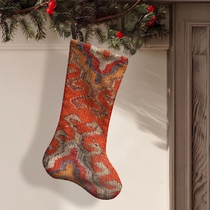 Könnte beinhalten: Ein Weihnachtsstrumpf mit einem gewebten Textilmuster in den Farben gebranntes Orange, tiefes Rot, gedämpftes Blau und Creme. Der Strumpf hat eine strukturierte, teppichartige Optik mit einem geometrischen Muster und einer schwarzen Aufhängeschlaufe.