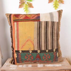 May include: Square decorative pillow with a patchwork design. The pillow features a variety of colors and patterns, including yellow, red, black, and green. The pillow is made of a textured fabric and has a brown border.