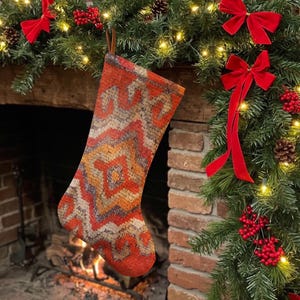 Könnte beinhalten: Ein Weihnachtsstrumpf mit einem gewebten geometrischen Muster in Orange-, Rot-, Grau- und Cremetönen. Der Strumpf hat eine braune Lederschlaufe zum Aufhängen an der Oberseite und besteht aus einem strukturierten Stoff mit sichtbarem Gewebe.
