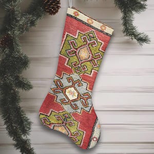 Könnte beinhalten: Ein Weihnachtsstrumpf mit rotem Hintergrund und einem geometrischen Muster in Grün, Grau und Gelb. Der Strumpf hängt an einer silbernen Schlaufe, mit einem Tannenzweig und Ornamenten im Hintergrund.
