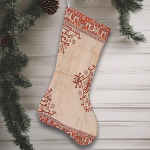 May include: Cream-colored Christmas stocking with a red and cream geometric pattern. The stocking is hanging from a white ribbon attached to a Christmas tree branch. The design features intricate details and a vintage aesthetic.