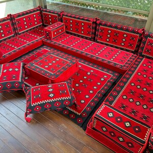 May include: A modular floor seating set featuring long rectangular cushions, backrests, and square floor pillows. The fabric is red and black with a repeating geometric pattern and small tassels on the corners of the cushions and backrests.