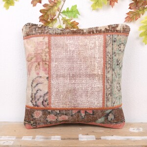 Op de afbeelding: Vierkant decoratief kussen met een patchwork ontwerp. Het kussen heeft een centraal lichtroze paneel, geflankeerd door patronen in bruin, groen en roze. Het is geplaatst op een houten oppervlak.