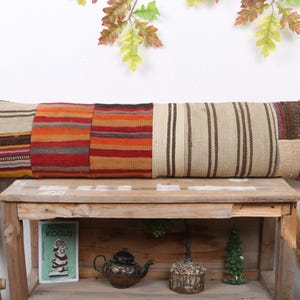 May include: A long, rectangular pillow with a patchwork design. The pillow features sections of various colors and patterns, including stripes and woven textures. It rests on a wooden table, showcasing its rustic aesthetic.
