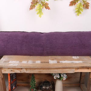Op de afbeelding: Een lang, rechthoekig paars kussen met een textuuroppervlak. Het kussen ligt op een houten bank. Decoratieve elementen zijn een kleine kerstboom, een theepot en een vaas met bloemen.