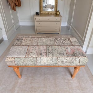May include: A rectangular ottoman featuring a patchwork upholstery design with muted tones of beige, gray, and faded pink. The top surface displays a variety of intricate, weathered patterns. The piece is supported by four light-colored wooden legs.