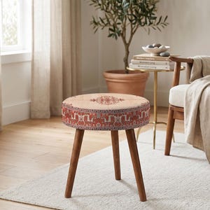 Può includere: Un pouf rotondo con piano beige e bordo a fantasia rosso e beige. Ha tre gambe angolate in legno marrone scuro. Il pouf è posto su un tappeto chiaro in una stanza con una palette di colori neutri.