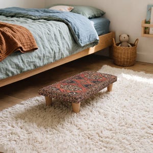 May include: A low rectangular footstool featuring a top surface upholstered in a patterned fabric with red, black, and cream geometric motifs. The stool is supported by four short, light-colored wooden legs with a smooth, cylindrical finish.