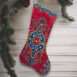Könnte beinhalten: Ein roter Weihnachtsstrumpf mit einem detaillierten blauen und grünen Muster. Der Strumpf hängt an einer weißen Schnur, mit einem Weihnachtsbaumzweig und Ornamenten im Hintergrund. Das Muster erinnert an ein traditionelles Teppichdesign.