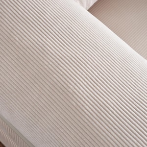 May include: Close-up of a light beige corduroy sofa. The fabric has a ribbed texture, with parallel lines running across the surface. The sofa appears to be part of a larger set, with a portion of another cushion visible.